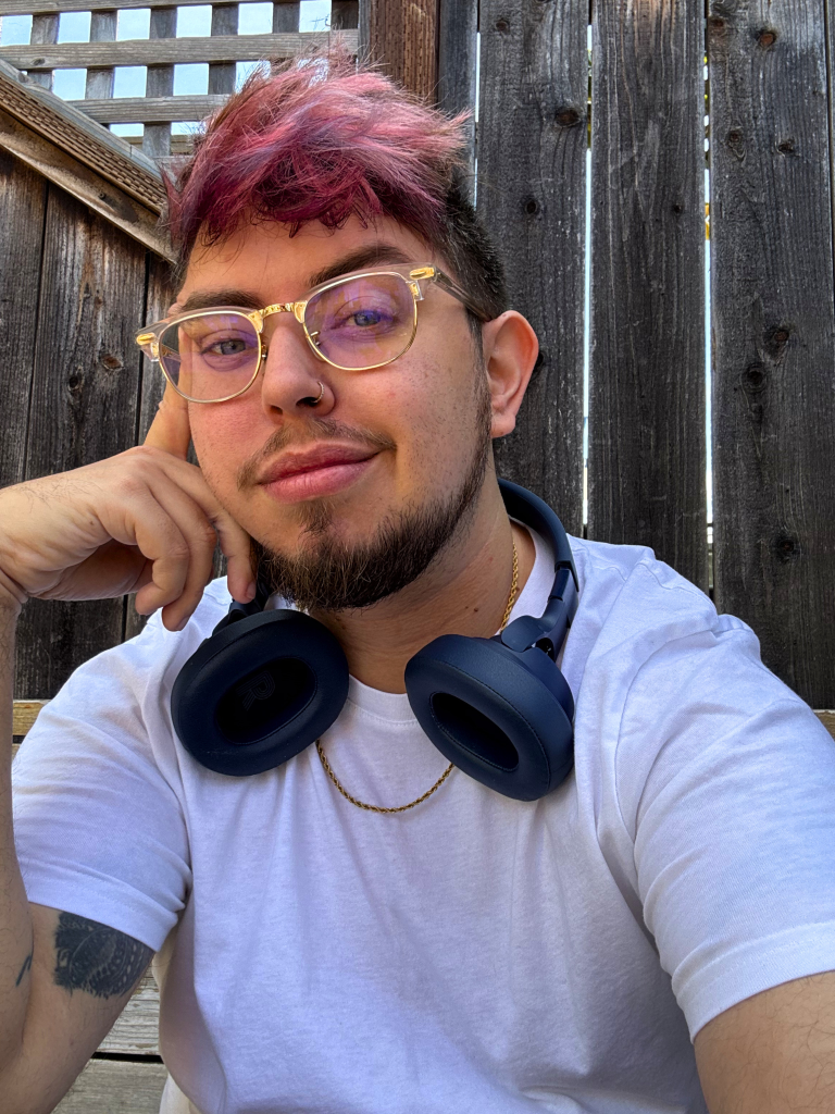Sam, a Maltese American transgender man with an olive complexion, gazes confidently into the camera. His hair is cut short with varying tones of pink, blush, and lavender. He has a pair of navy headphones around his neck and a gold chain against a crisp white tee. He is leaning his face against his index finger, smiling slightly. Behind him is a wooden fence (he is sitting on a cafe patio).