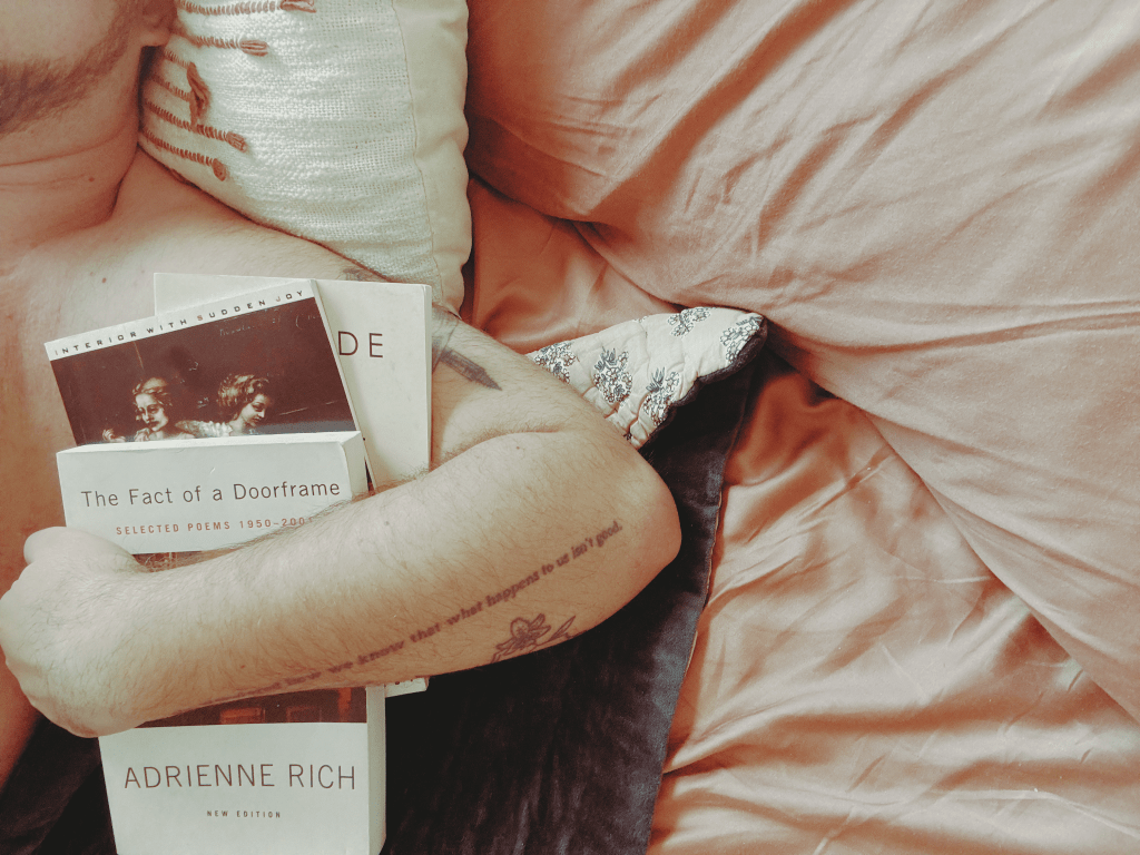A photo with a vintage filter. It features Sam's tattooed arm holding three books of poetry close to his chest, against a backdrop of teal and clay colored blankets and sheets.
